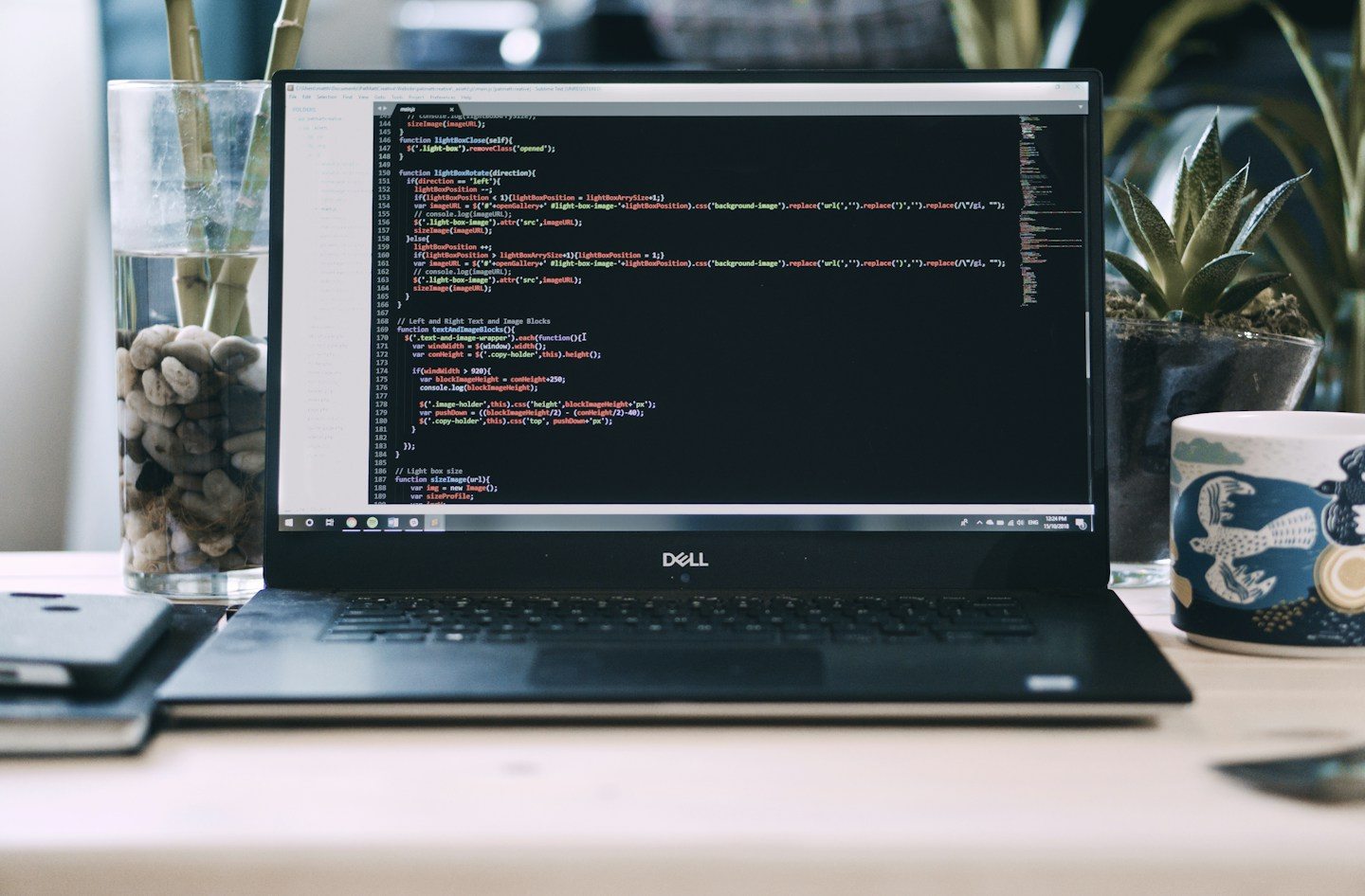 A laptop displaying website code on a desk beside a plant and a mug