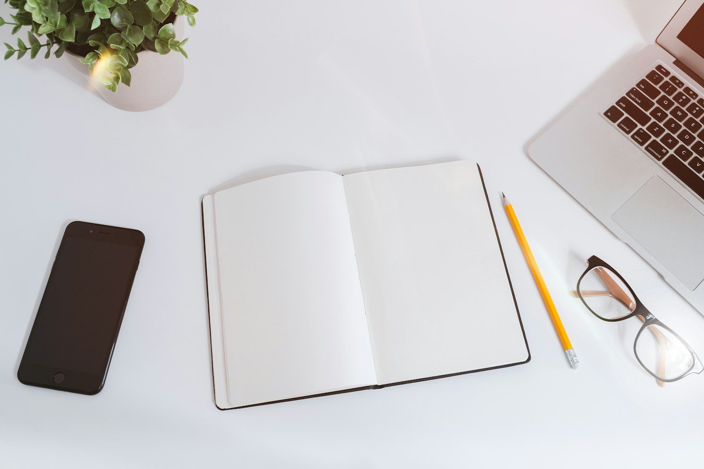 An open blank notebook, a phone, glasses and a laptop on a white desk