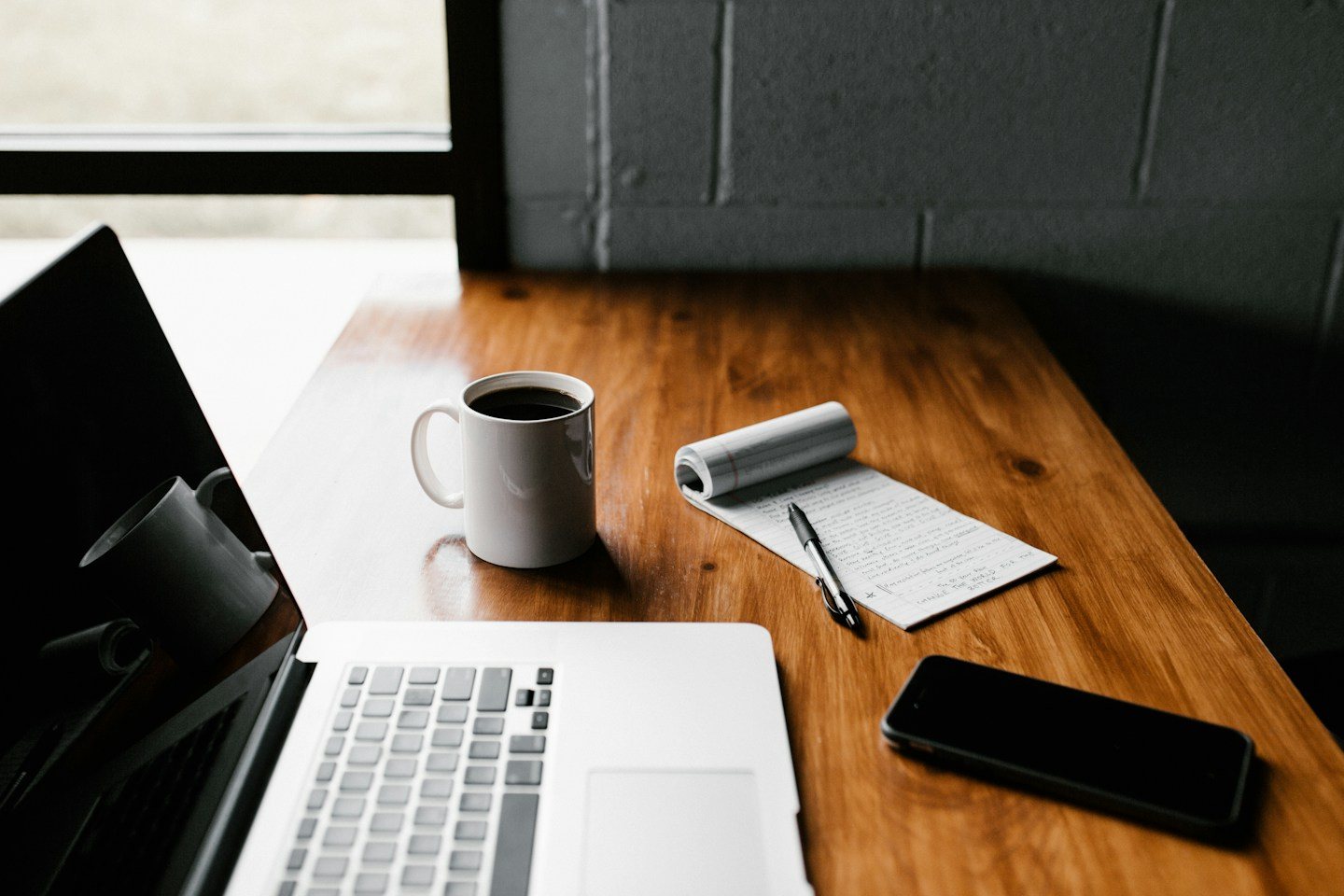 A laptop, handwritten notes, a mug and a phone on a wooden desk
