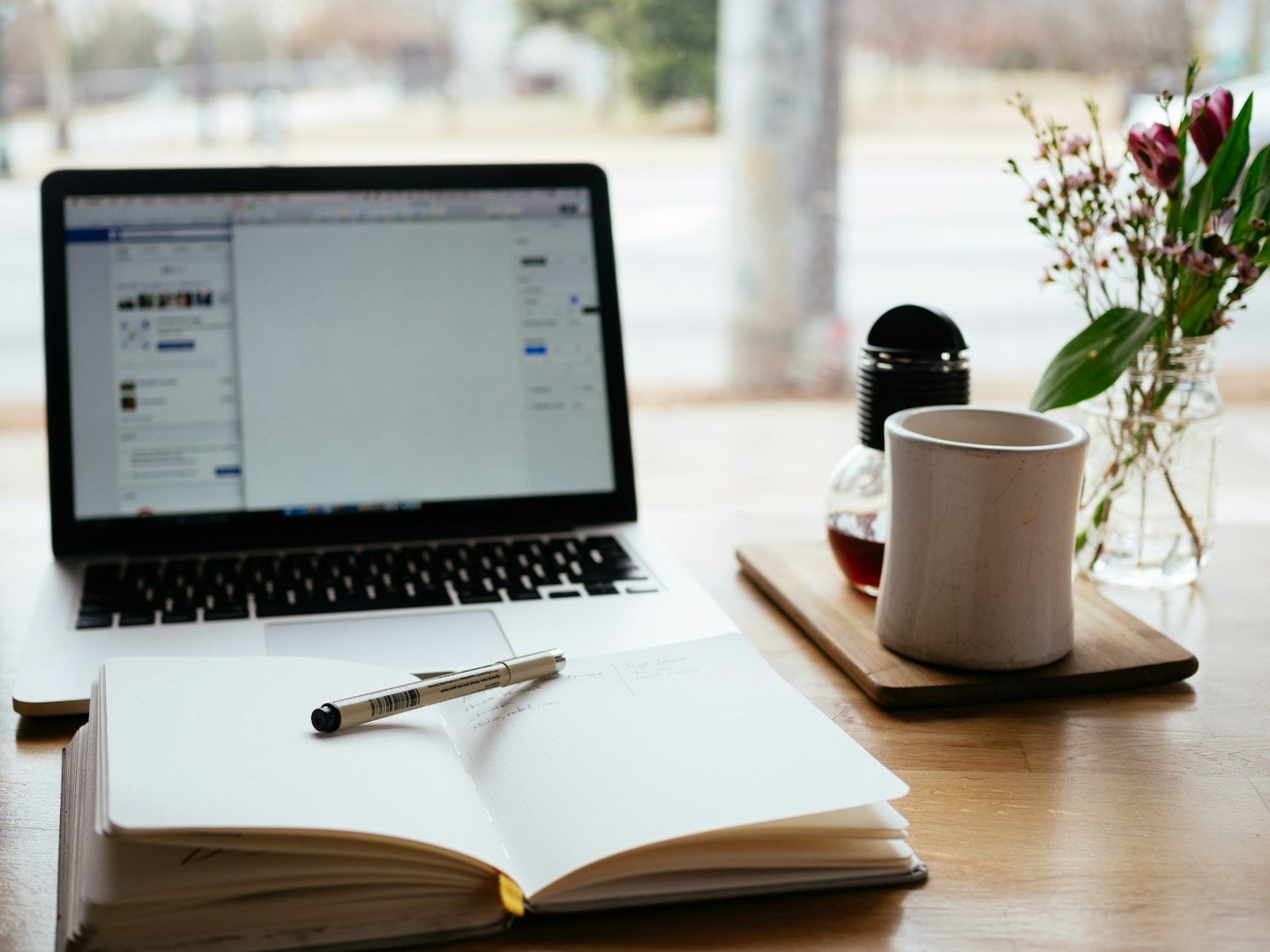 An open notebook and pen beside a laptop and mug at a desk